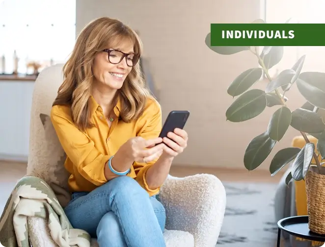 Middle-aged woman wearing glasses sitting in a chair looking at phone and smiling with "individuals" text tag